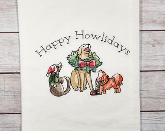 Op de afbeelding: Witte keukendoek met geborduurde tekst "Happy Howlidays" en drie honden in kerstkledij. De hond in het midden draagt een krans om zijn nek.