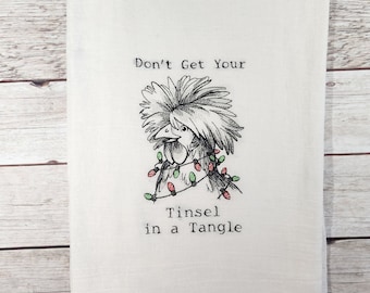 Op de afbeelding: Een witte keukendoek met een zwart-witte illustratie van een haan met rode en groene kerstverlichting om zijn kop gewikkeld. De tekst "Don't Get Your Tinsel in a Tangle" staat onder de illustratie.