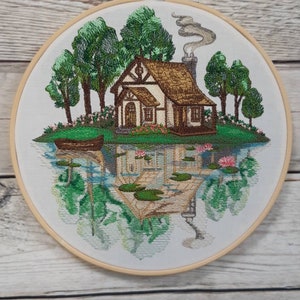 May include: Embroidered hoop art depicting a cottage with a brown roof and white walls, surrounded by green trees and a lake with lily pads. The cottage has a chimney with smoke coming out of it. The scene is reflected in the water.