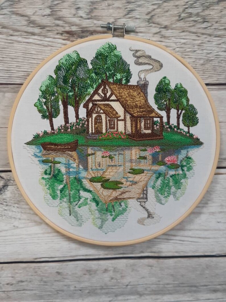 May include: Embroidered hoop art depicting a cottage with a brown roof and white walls, surrounded by green trees and a lake with lily pads. The cottage has a chimney with smoke coming out of it. The scene is reflected in the water.