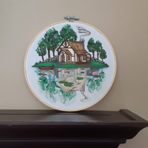 May include: Embroidered hoop art depicting a cottage with a brown roof and white walls, surrounded by green trees and a reflection of the scene in a body of water with lily pads.
