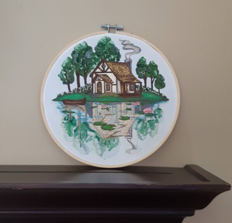 May include: Embroidered hoop art depicting a cottage with a brown roof and white walls, surrounded by green trees and a reflection of the scene in a body of water with lily pads.