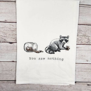 K&ouml;nnte beinhalten: Ein wei&szlig;es Geschirrtuch mit einem gestickten Bild eines Waschb&auml;ren, der ein Keks h&auml;lt, und einer Dose Kekse auf dem Boden. Der Text "You saw nothing" ist unter dem Bild gestickt.