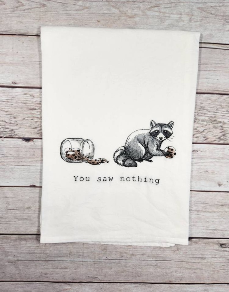 K&ouml;nnte beinhalten: Ein wei&szlig;es Geschirrtuch mit einem gestickten Bild eines Waschb&auml;ren, der ein Keks h&auml;lt, und einer Dose Kekse auf dem Boden. Der Text "You saw nothing" ist unter dem Bild gestickt.