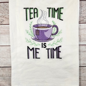 Könnte beinhalten: Weißes Geschirrtuch mit gesticktem Text "Tea Time is Me Time" und einer lila Teetasse mit Dampf, der aus ihr aufsteigt.