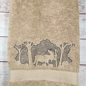May include: A tan-coloured terry cloth towel featuring an embroidered design of a moose standing among trees. The embroidery uses dark brown thread to create a silhouette effect against the textured fabric of the towel.