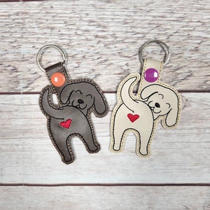 May include: Two dog-shaped keyrings with a heart on the back. One keyring is brown leather with an orange key ring and the other is beige leather with a purple key ring.