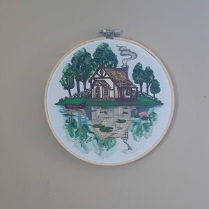 May include: Embroidered hoop art featuring a cottage with a chimney and smoke, surrounded by trees and water lilies. The cottage is reflected in the water.