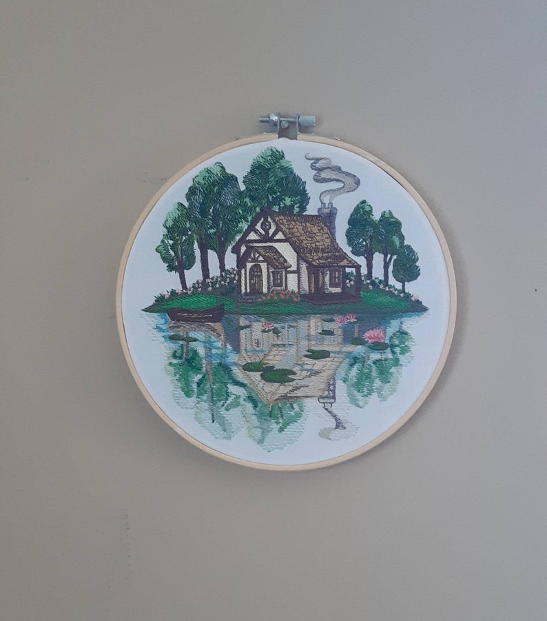 May include: Embroidered hoop art featuring a cottage with a chimney and smoke, surrounded by trees and water lilies. The cottage is reflected in the water.
