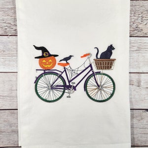 Könnte beinhalten: Ein lila Fahrrad mit einem Kürbis, einem schwarzen Raben und einer schwarzen Katze im Korb. Das Fahrrad hat ein Spinnennetz-Design am Rahmen.