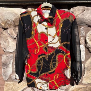 May include: A vintage button-down shirt with a bold chain and rope pattern in gold, black, red, and white. The shirt has sheer black sleeves and a black collar. The label reads "Joy Jacobs".