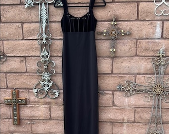 May include: A black, floor-length dress with a velvet bodice and spaghetti straps. The bodice features vertical lines and decorative accents. The dress hangs on a black hanger against a brick wall.