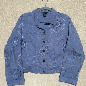 May include: A light blue denim jacket with embroidered white and blue floral designs. The jacket has a button closure and long sleeves.