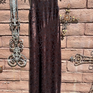 May include: A sleeveless floor-length dress in a deep brown hue, featuring a tonal floral pattern throughout the fabric. The garment has a round neckline and a straight, column-like silhouette, hanging from a black hanger against a brick wall.