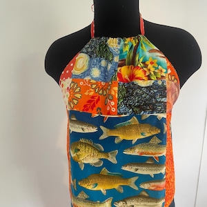 Op de afbeelding: Een patchwork haltertop met diverse patronen. De top heeft een blauw paneel met visillustraties. Andere panelen tonen bloemen- en artistieke ontwerpen in oranje, blauw en geel. De halterbandjes zijn rood.