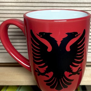 Może przedstawiać: Czerwony kubek ceramiczny z czarnym wzorem dwugłowego orła. Orzeł jest symbolem narodowym Albanii.