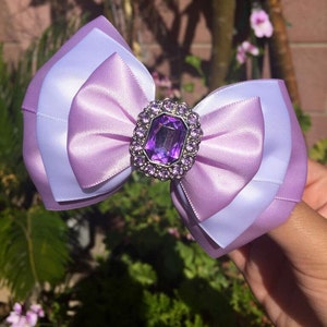 Puede incluir: Un gran lazo de satén morado y blanco con un gran strass morado en el centro. El lazo está decorado con strass más pequeños alrededor del borde.