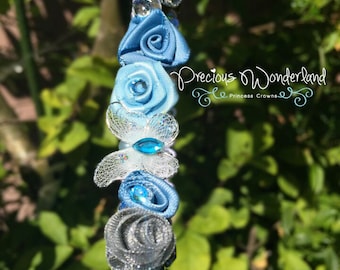 May include: A blue and silver headband with a variety of blue satin rosettes, a white butterfly with a blue gem, and silver glitter accents. The headband is decorated with the text "Precious Wonderland - Princess Crowns".