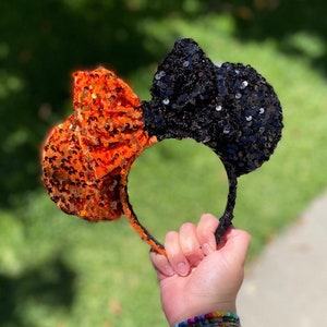 May include: A Halloween-themed headband with two sequined bows. One bow is orange, the other is black. The headband is held up against a blurred green background.