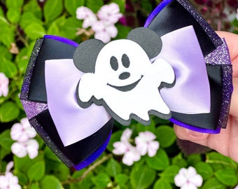 Puede incluir: Un lazo para el pelo con temática de Halloween con un fantasma blanco sonriente y orejas de Mickey Mouse. El lazo tiene cintas negras, moradas y lavanda con detalles de purpurina. Fondo de hojas verdes y pequeñas flores blancas.