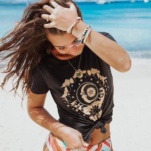 May include: A woman wearing a black t-shirt with a gold moon and stars graphic and a colourful striped bikini bottom. She is standing on a white sandy beach with blue water in the background.