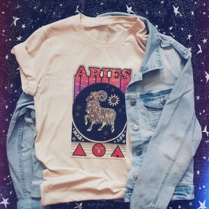 May include: A beige t-shirt with a vintage-style graphic of the zodiac sign Aries. The graphic features a ram with a sun and moon, and the text "Aries" in a retro font. The dates "Mar. 21" and "Apr. 19" are also included in the graphic.