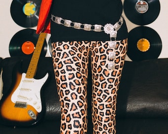 May include: A woman wearing a red blazer over a black t-shirt with a graphic print of a skull and feathers. The t-shirt also has the text "Valley 1994". She is wearing leopard print leggings and a silver belt with a large buckle. The background features a wall with a collection of vinyl records.