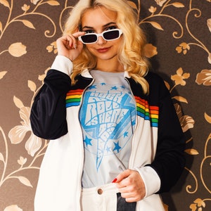 May include: A white jacket with black sleeves and a rainbow stripe detail. The jacket is worn over a light blue t-shirt with the text "Rock Roll is Here" printed on it. The person is wearing white trousers with a black flame design.