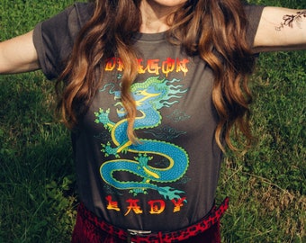 May include: A woman wearing a dark grey t-shirt with a green and blue dragon graphic and the text "Dragon Lady". She is wearing a red velvet skirt with a leopard print belt.