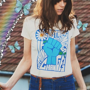 May include: A white t-shirt featuring a blue and white graphic print of a fist with the text "Nothing Changes If Nothing Changes". The shirt is being worn by a person with long brown hair and a brown leather belt.