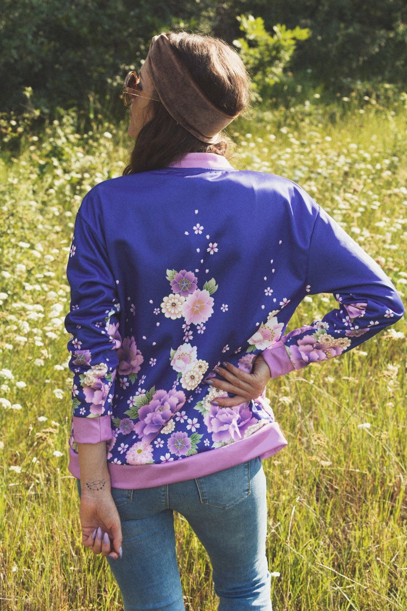 May include: A purple bomber jacket with a floral print featuring pink, white, and blue flowers. The jacket has a pink ribbed collar and cuffs.