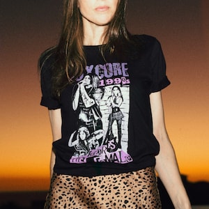 May include: A woman wearing a black t-shirt with a graphic print of a band called "Joycore 1990s" and a brown leopard print skirt.