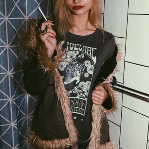 Peut inclure: Une femme portant une veste en fausse fourrure brune avec un t-shirt noir orn&eacute; d'un motif graphique vintage qui dit "Bluefire Fest '7968". Elle porte une jupe &agrave; motifs noir et blanc et a de longs cheveux blonds.