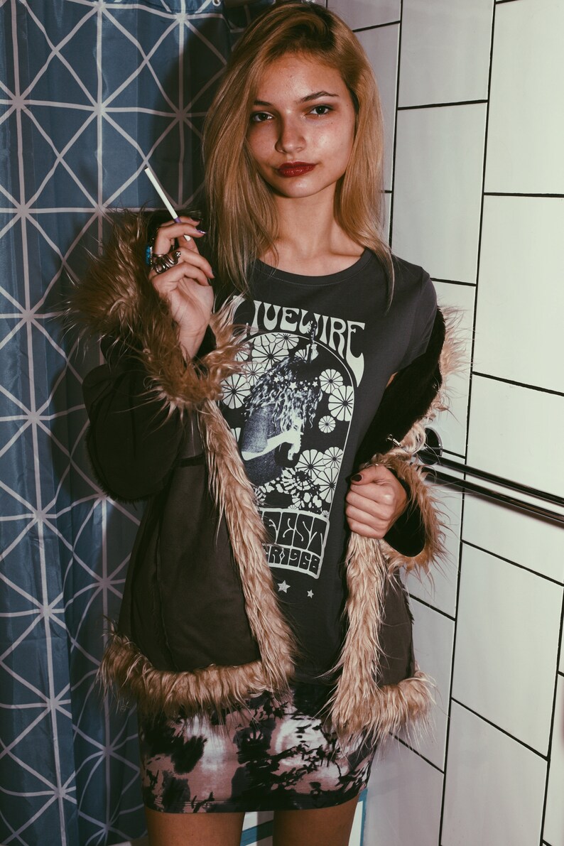 Peut inclure: Une femme portant une veste en fausse fourrure brune avec un t-shirt noir orn&eacute; d'un motif graphique vintage qui dit "Bluefire Fest '7968". Elle porte une jupe &agrave; motifs noir et blanc et a de longs cheveux blonds.