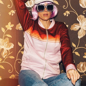 May include: A woman wearing a pink and red striped jacket with a white visor and large square sunglasses. She is sitting on a brown couch with floral wallpaper.