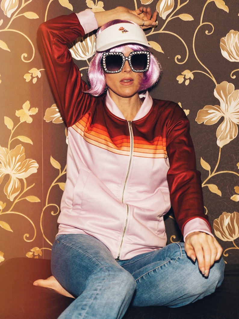 May include: A woman wearing a pink and red striped jacket with a white visor and large square sunglasses. She is sitting on a brown couch with floral wallpaper.