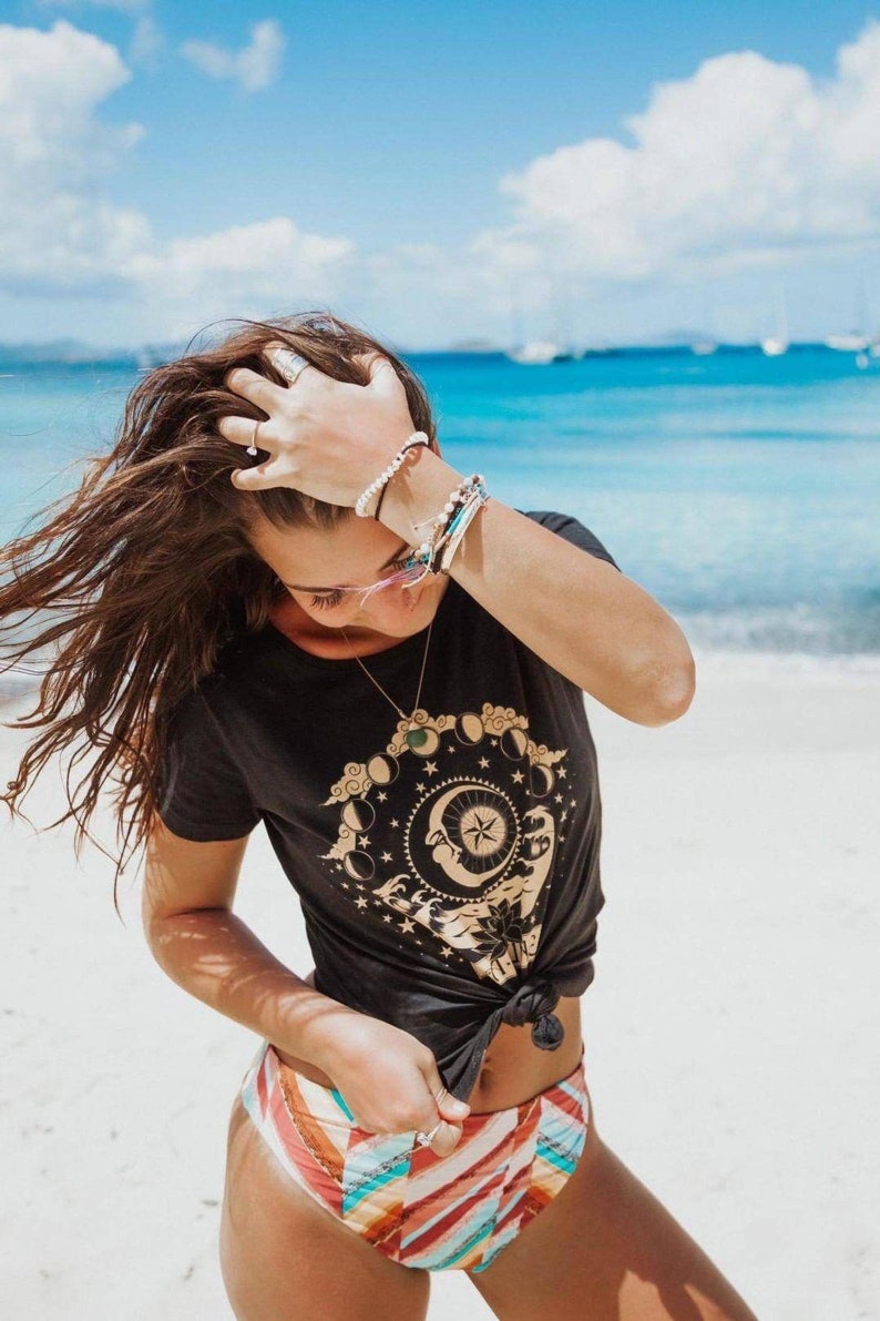 May include: A woman wearing a black t-shirt with a gold moon and stars graphic and a colourful striped bikini bottom. She is standing on a white sandy beach with blue water in the background.