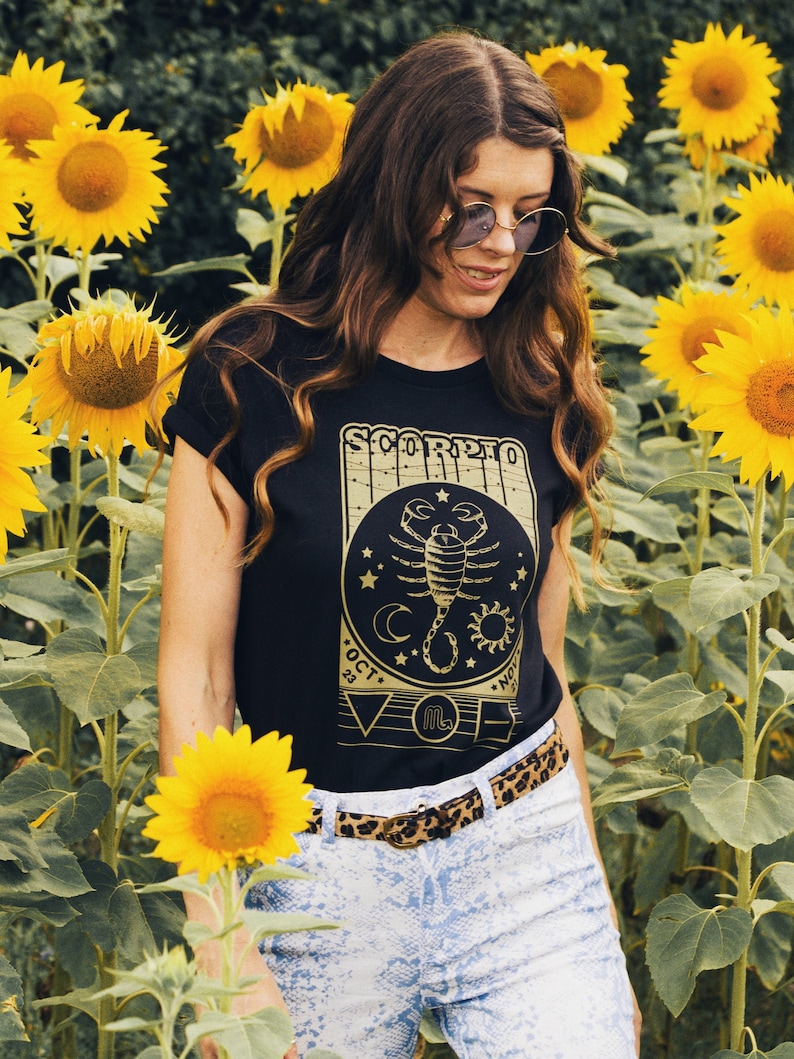 May include: A woman wearing a black t-shirt with a gold scorpion graphic and the text "Scorpio Oct 23 - Nov 21" and a leopard print belt. She is standing in a field of sunflowers.