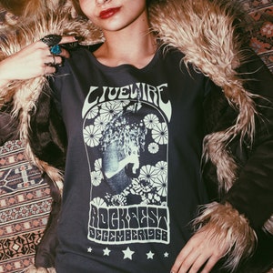 Peut inclure: Une femme portant une chemise grise &agrave; manches longues avec un imprim&eacute; graphique blanc qui dit "Livewire Rockfest December 1968". Elle porte une veste en fausse fourrure brune et une jupe en tie-dye noir et blanc.