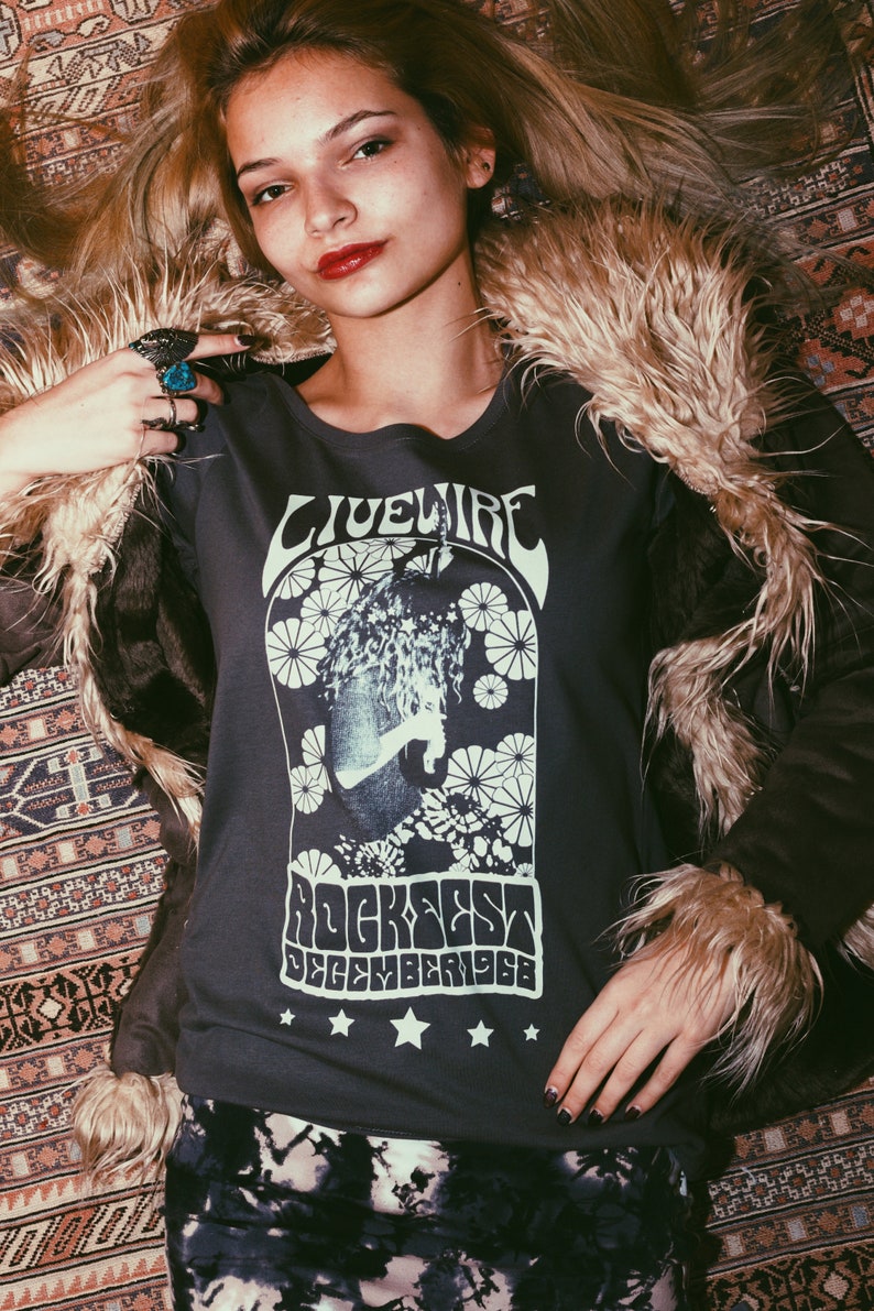 Peut inclure: Une femme portant une chemise grise &agrave; manches longues avec un imprim&eacute; graphique blanc qui dit "Livewire Rockfest December 1968". Elle porte une veste en fausse fourrure brune et une jupe en tie-dye noir et blanc.