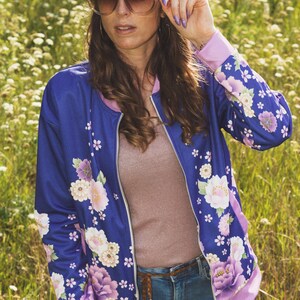 May include: A woman wearing a blue floral bomber jacket with a pink lining, brown headband, and sunglasses. She is standing in a field of purple flowers.
