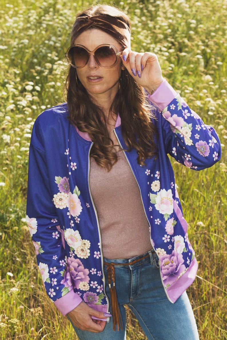 May include: A woman wearing a blue floral bomber jacket with a pink lining, brown headband, and sunglasses. She is standing in a field of purple flowers.