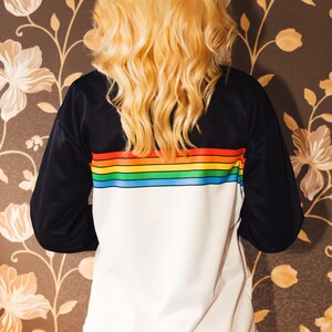 May include: A white sweatshirt with black sleeves and a rainbow stripe across the chest. The sweatshirt is being worn by a person with long blonde hair.