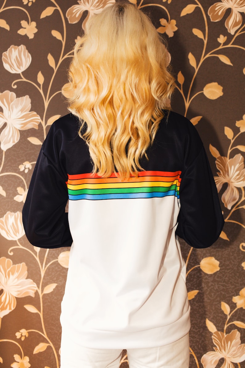 May include: A white sweatshirt with black sleeves and a rainbow stripe across the chest. The sweatshirt is being worn by a person with long blonde hair.