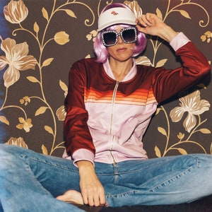 May include: A person wearing a pink and red striped jacket, blue jeans, a white visor with a pink flower design, and pink sunglasses. The person is sitting on a couch with a floral patterned background.