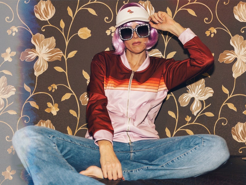 May include: A person wearing a pink and red striped jacket, blue jeans, a white visor with a pink flower design, and pink sunglasses. The person is sitting on a couch with a floral patterned background.