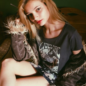 Peut inclure: Une femme portant un t-shirt gris avec le texte "Livewire" imprim&eacute; dessus. Elle porte &eacute;galement une veste en fausse fourrure brune et une jupe &agrave; motifs.