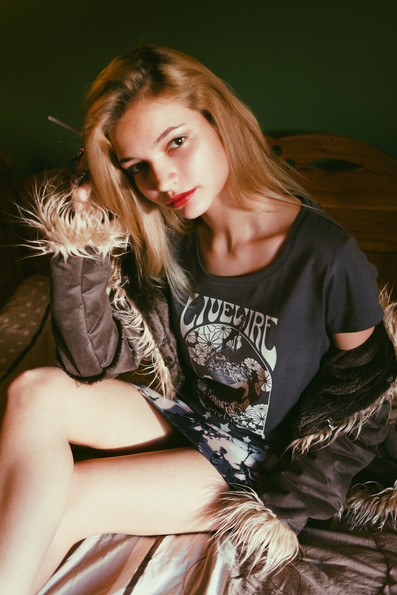 Peut inclure: Une femme portant un t-shirt gris avec le texte "Livewire" imprim&eacute; dessus. Elle porte &eacute;galement une veste en fausse fourrure brune et une jupe &agrave; motifs.