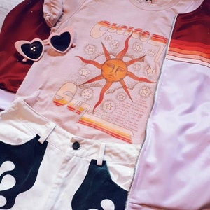 May include: A light brown t-shirt with a graphic of a sun with a face and the word "Sun" in a retro font. The shirt is layered over a red jacket with a purple and white striped sleeve. A pair of pink heart-shaped sunglasses are on top of the shirt. White denim shorts with a black flame design are visible in the bottom of the image.