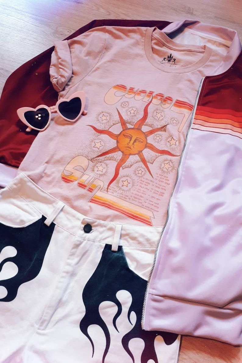 May include: A light brown t-shirt with a graphic of a sun with a face and the word "Sun" in a retro font. The shirt is layered over a red jacket with a purple and white striped sleeve. A pair of pink heart-shaped sunglasses are on top of the shirt. White denim shorts with a black flame design are visible in the bottom of the image.